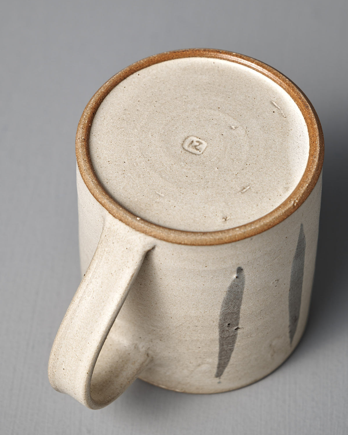 The Mug – Stripes by Zoë Isaacs is a handmade ceramic mug featuring two vertical brushstrokes, a matte cream glaze, and a circular stamp on its unglazed, iron-rich clay base.