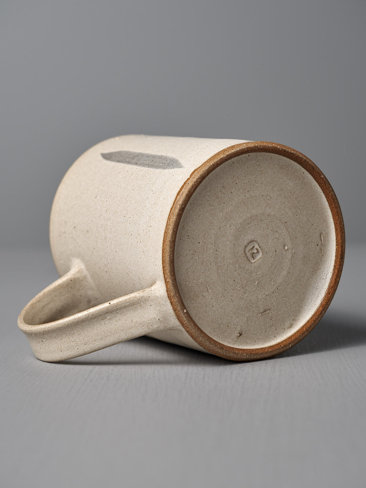 The Mug – Stripes by Zoë Isaacs, a handmade ceramic mug with a matte cream glaze, rests on its side to reveal its iron-rich clay base stamped with the maker’s mark and part of the handle.