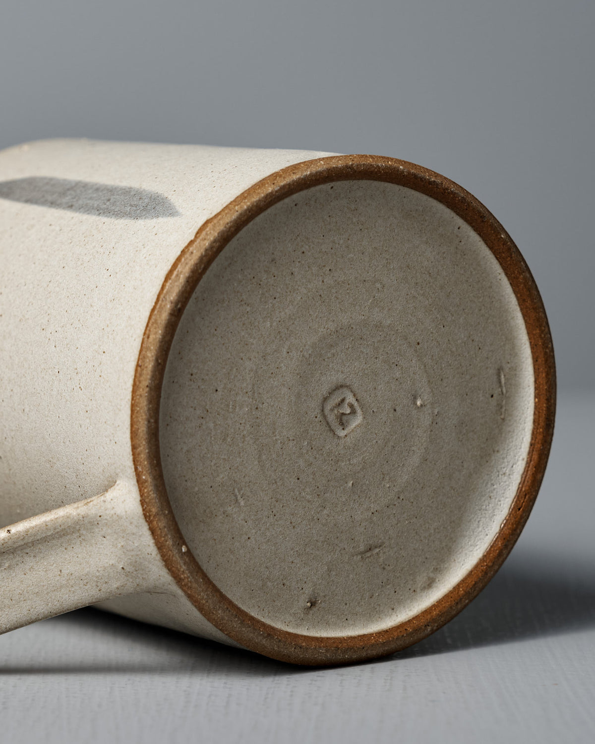 The Mug – Stripes by Zoë Isaacs, a handmade ceramic mug with a matte cream glaze, rests on its side to reveal its iron-rich clay base stamped with the maker’s mark and part of the handle.