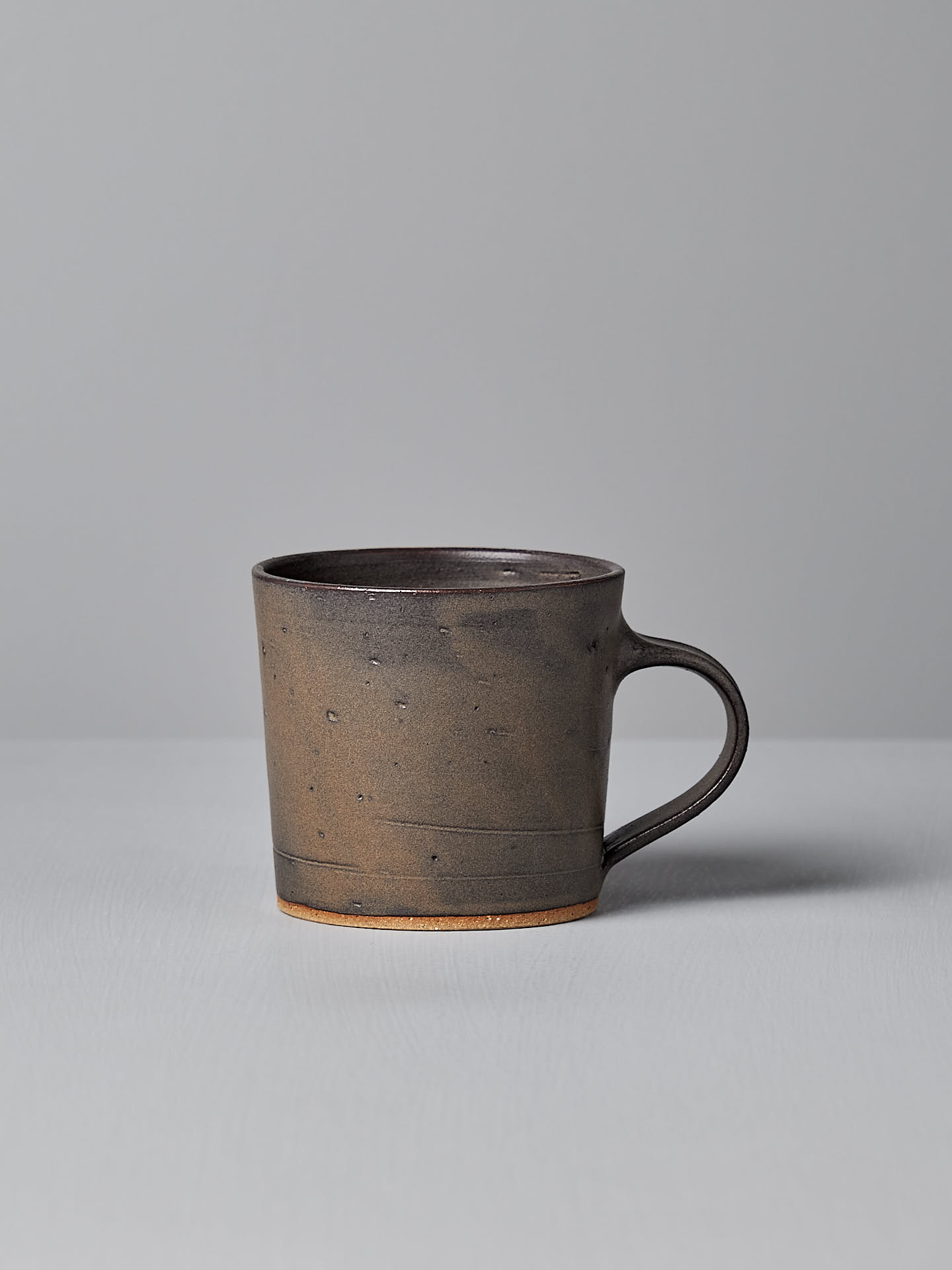 A Zoë Isaacs Coffee Cup – Dapple Brown, featuring a matte brown glaze and handle, sits on a light grey surface against a grey background.