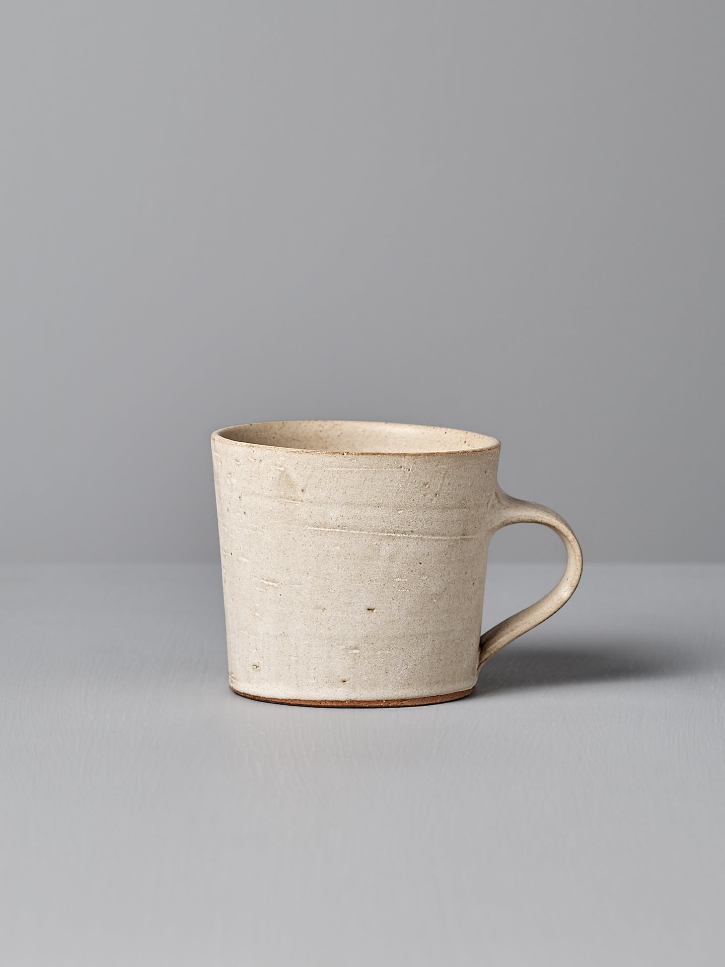 A Zoë Isaacs Coffee Cup – Seafoam, featuring a handle and a matte cream glaze, is placed on a light grey surface against a grey background.