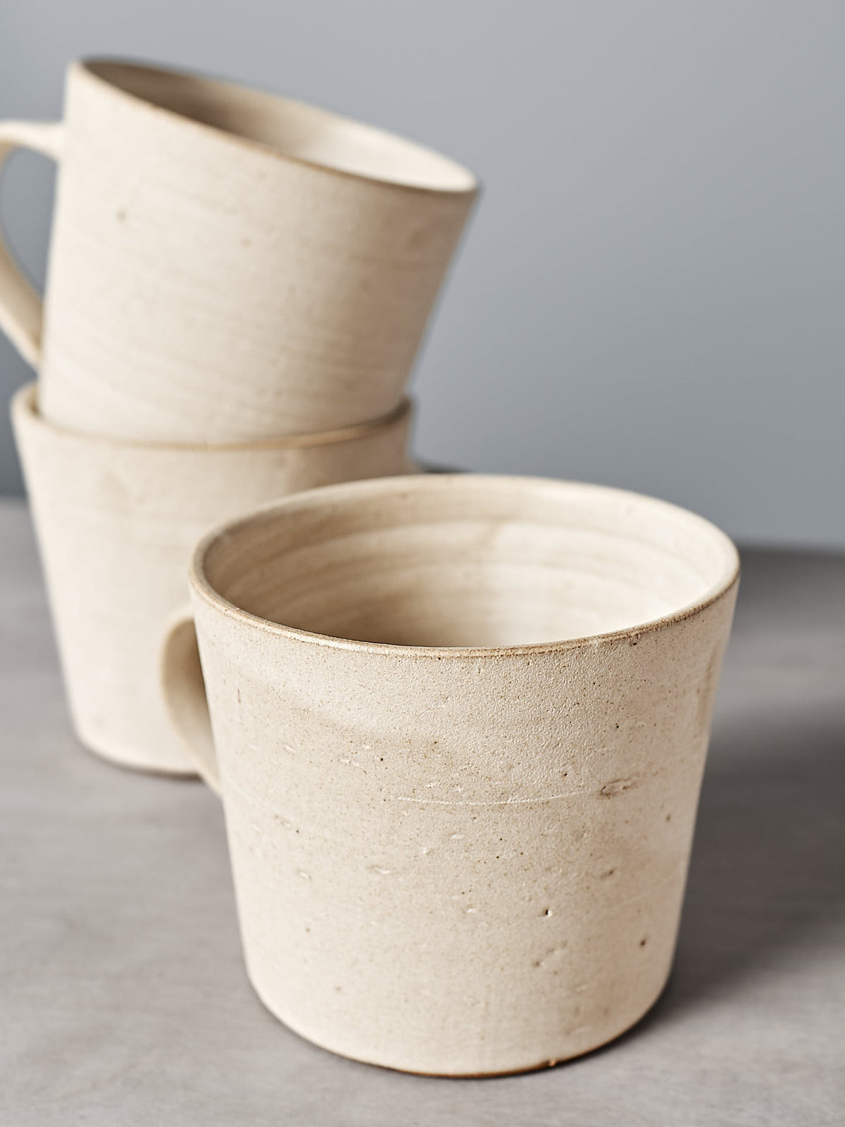 Three Zoë Isaacs Coffee Cups in seafoam are stacked on a gray surface, with one positioned in front and two behind. The handmade mugs boast a matte seafoam glaze that complements the plain gray wall backdrop.