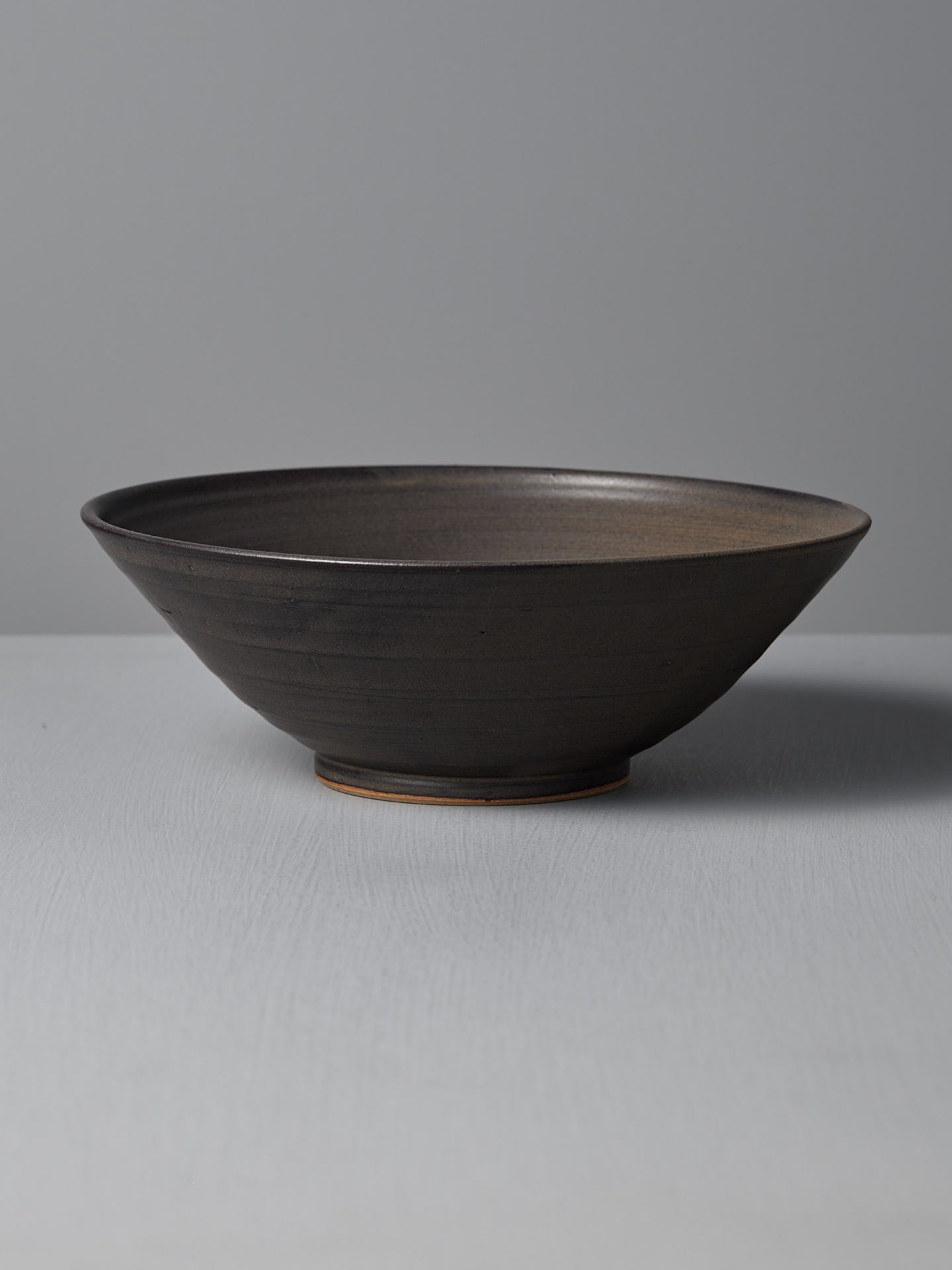 The Zoë Isaacs Serving Bowl – Dapple Brown is a dark handmade ceramic bowl with a wide rim and matte finish, displayed on a light gray surface against a plain background, making it an elegant decorative piece.