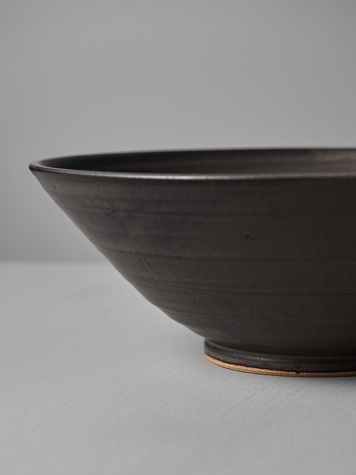 A close-up of the Zoë Isaacs Serving Bowl – Dapple Brown, featuring a dark ceramic matte finish, set on a light gray surface with a plain backdrop.