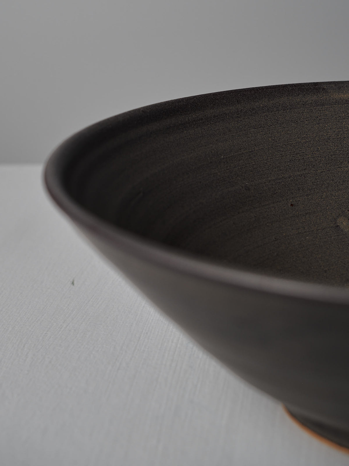 Close-up of the Zoë Isaacs Serving Bowl – Dapple Brown, a handmade dark ceramic piece, partially visible on a light gray surface.