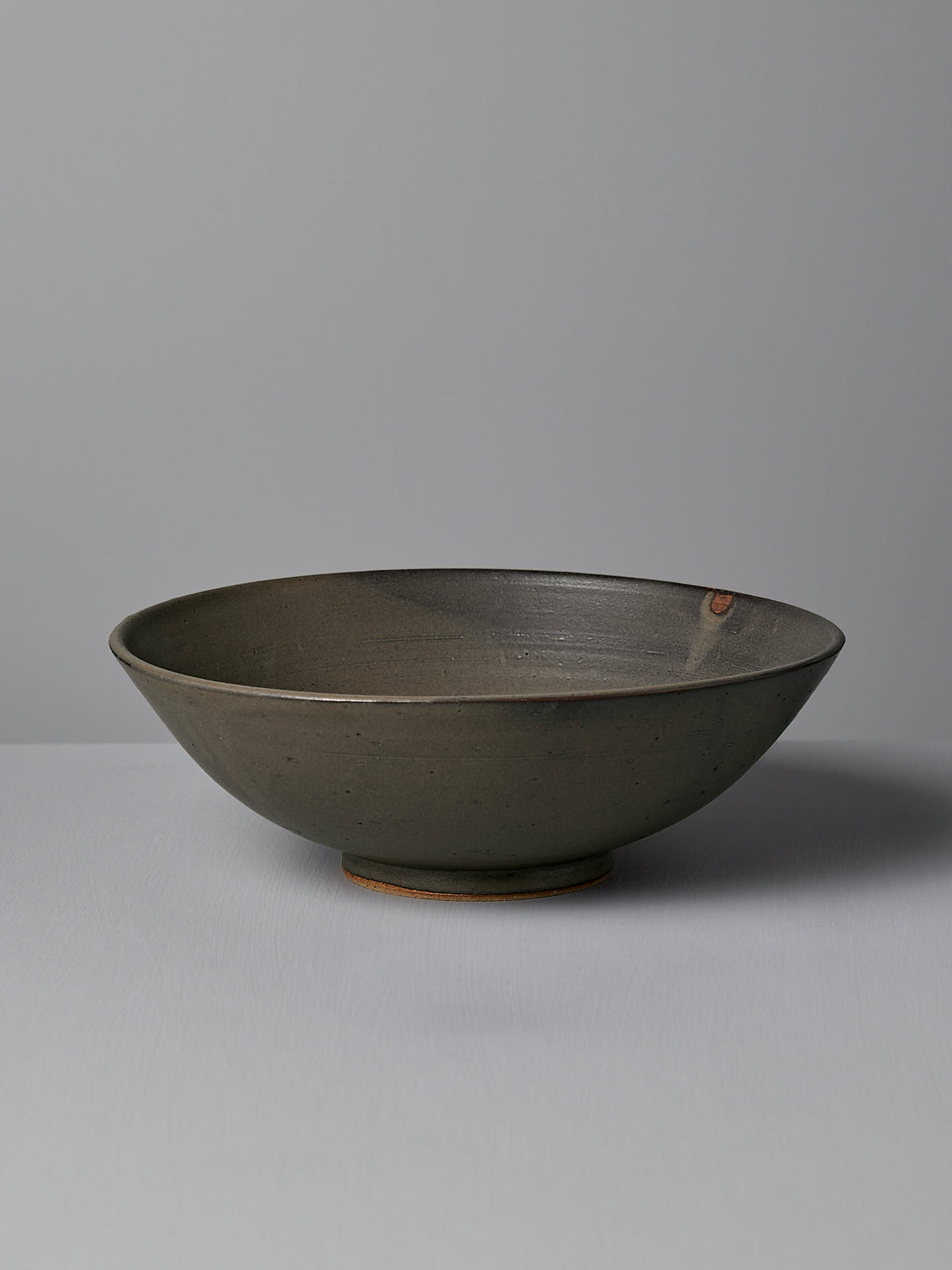 The Zoë Isaacs Serving Bowl – Dapple Brown, a shallow ceramic piece with a matte gray finish, wheel-thrown from toast-colored clay, featuring a slightly tapered rim and round base, sits on a light gray surface against a plain background.