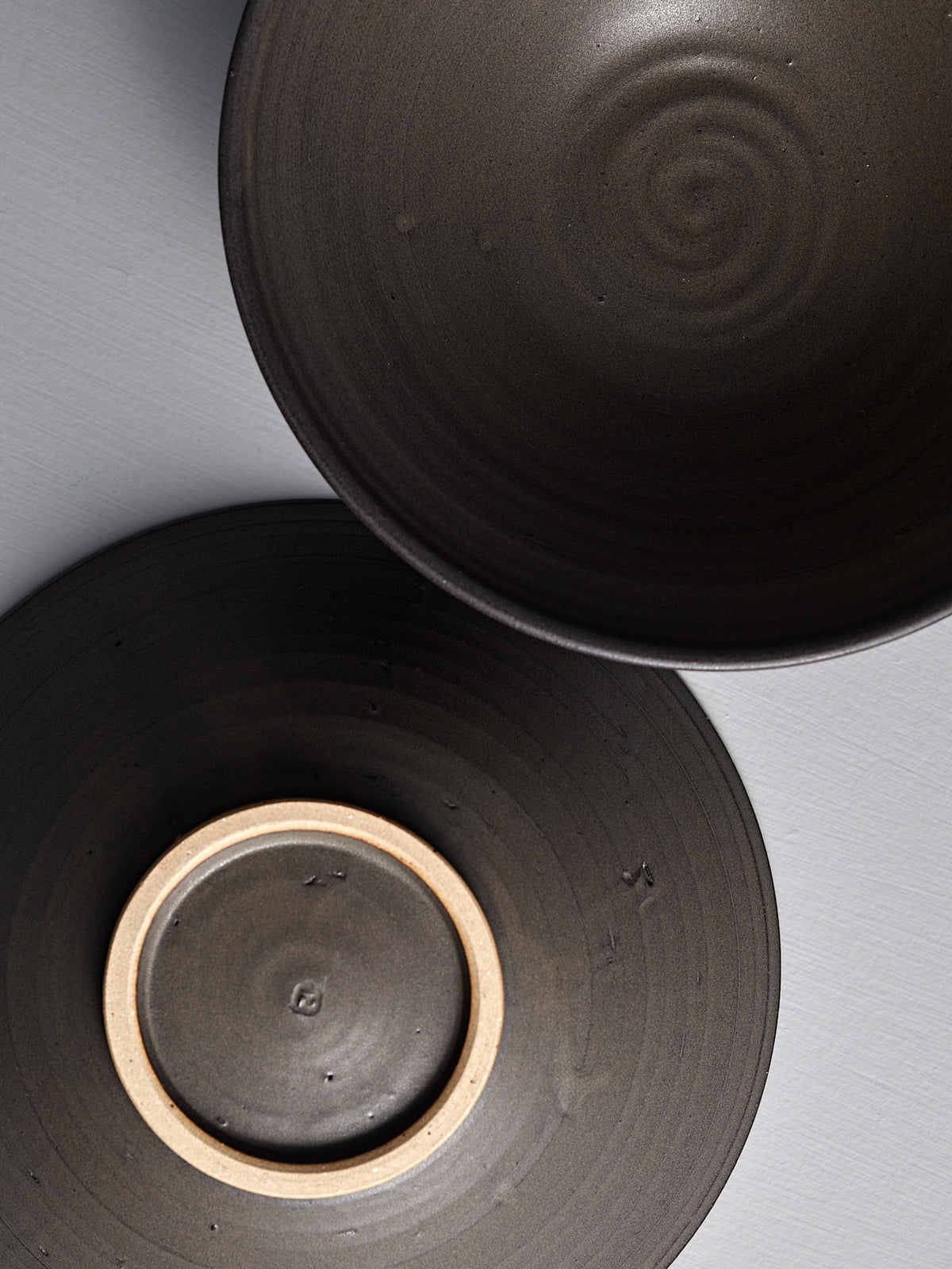 Two Zoë Isaacs “Dapple Brown” serving bowls—handmade dark ceramics with circular patterns, one upright showing its interior and the other upside down revealing a smooth base—make a unique decorative accent on a light gray surface.