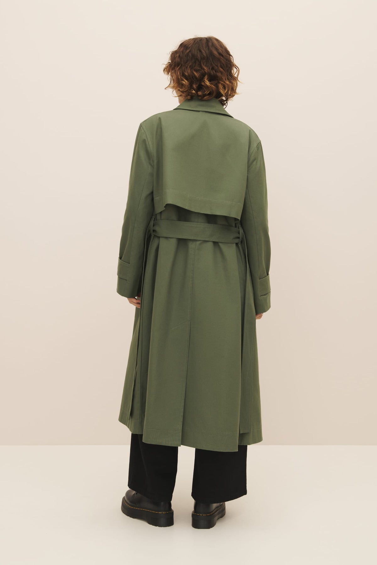 Woman standing with her back to the camera, wearing a Kowtow Cleo Trench Coat in Sage and black trousers in a relaxed fit.