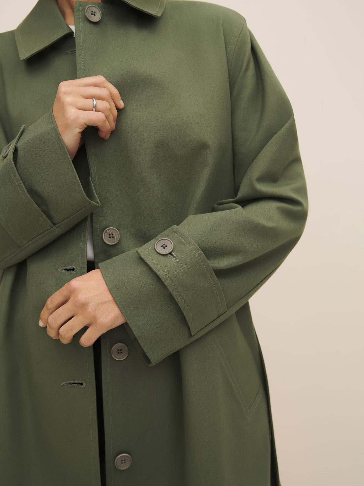 Person wearing a Kowtow Cleo Trench Coat – Sage with a relaxed fit and buttons, holding the lapel, showcasing the design and fabric.