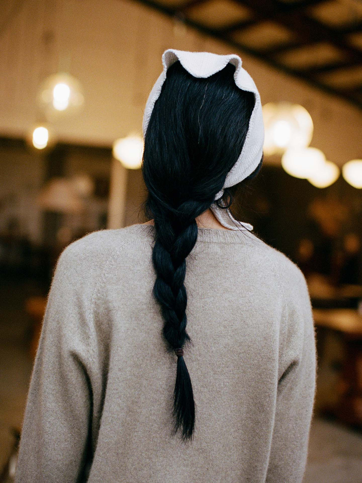A person with long black hair in a 97cm braid, wearing a beige sweater and the Daisy Scarf – Ivory, is shown from behind indoors under warm lighting.