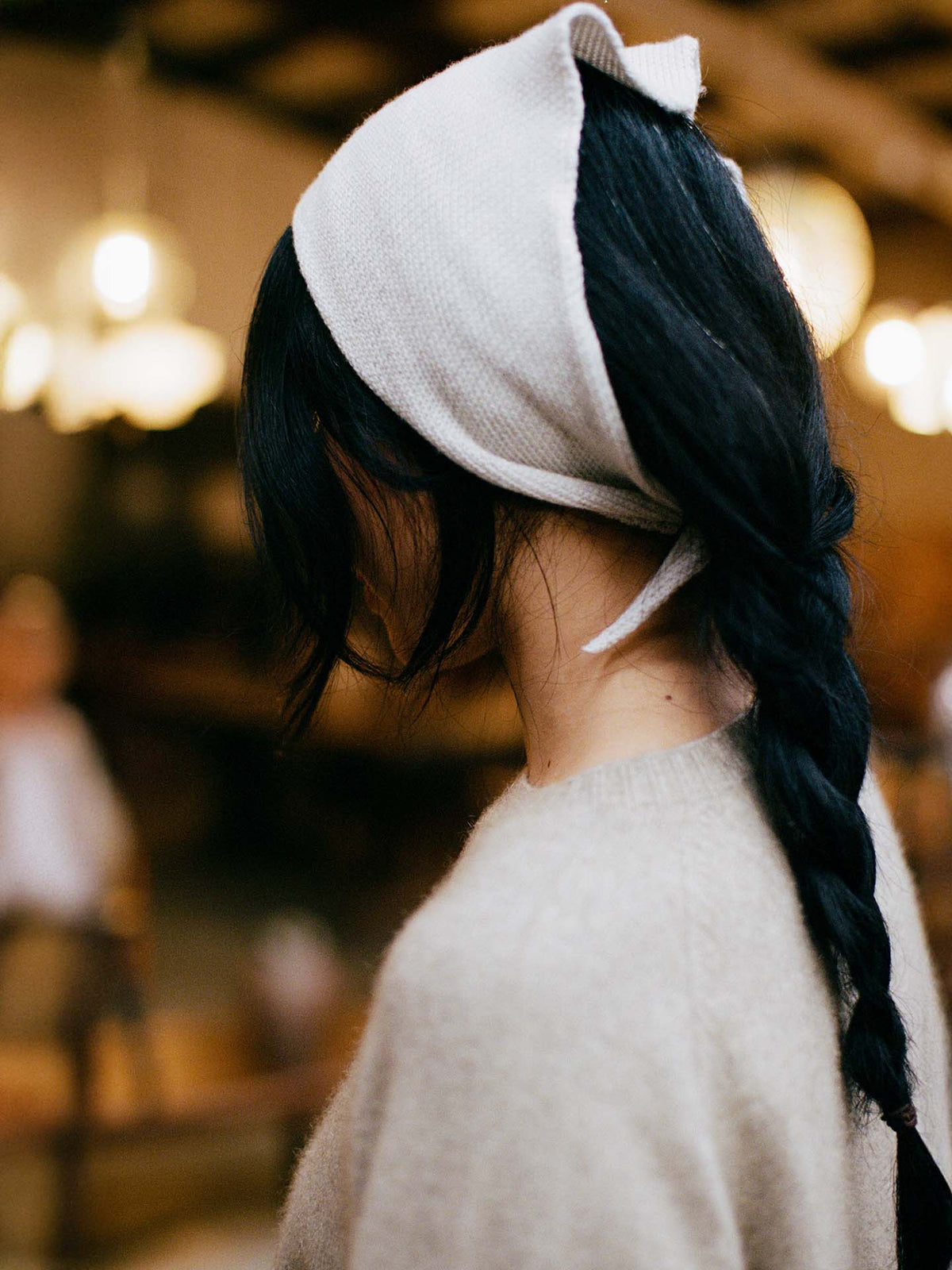 Person with long dark hair in a braid, wearing the Daisy Scarf – Ivory and a beige sweater, photographed from the side indoors; scarf dimensions: 97cm long, 22cm wide.