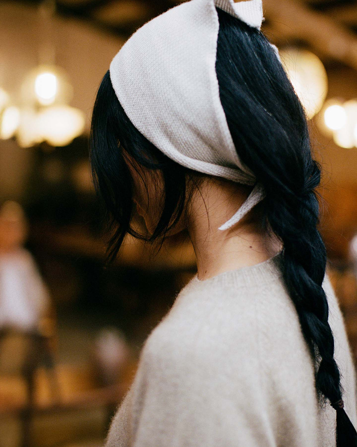 Person with long dark hair in a braid, wearing the Daisy Scarf – Ivory and a beige sweater, photographed from the side indoors; scarf dimensions: 97cm long, 22cm wide.