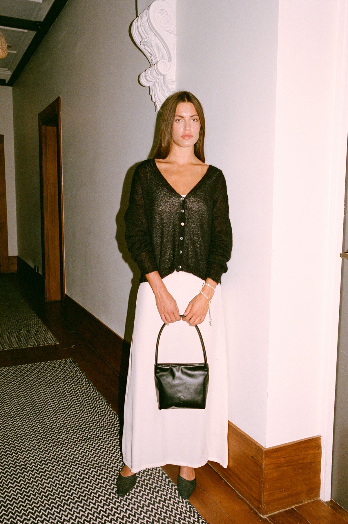 A petite woman stands by a white wall indoors, wearing the Mist Cardigan in black with a white skirt, black shoes, and holding a black handbag. The space features wooden trim and patterned flooring.