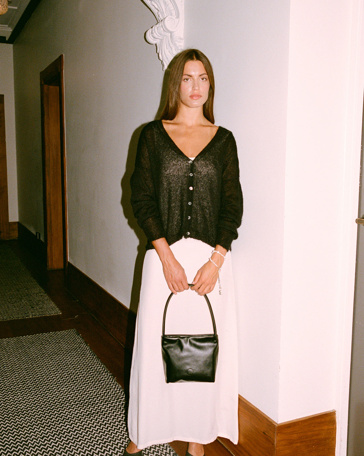 A petite woman stands by a white wall indoors, wearing the Mist Cardigan in black with a white skirt, black shoes, and holding a black handbag. The space features wooden trim and patterned flooring.