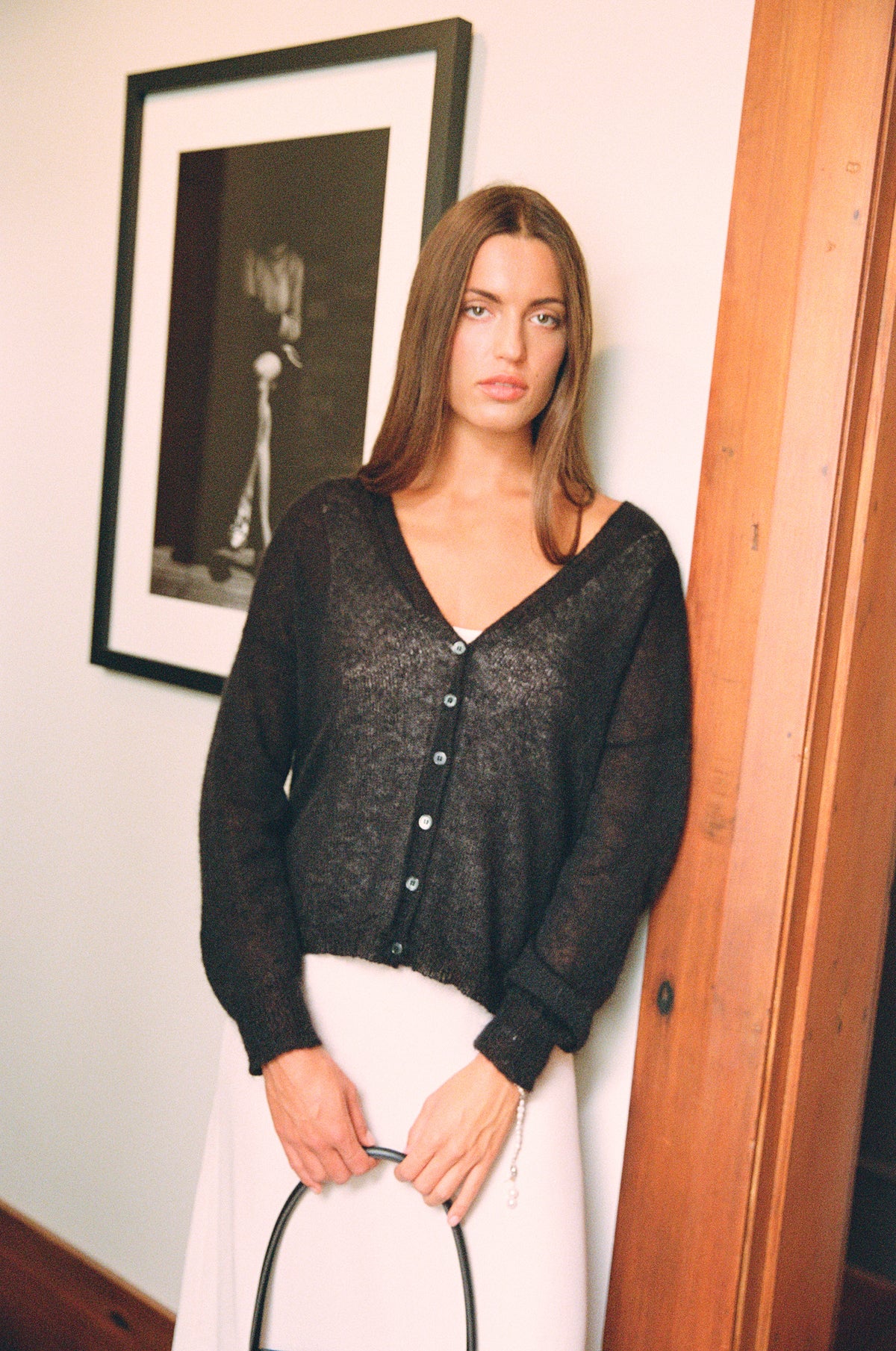 A petite woman with long brown hair stands indoors by a wooden panel, wearing the Mist Cardigan – Black and a light skirt, holding a black handle with both hands. A framed photo hangs behind her.