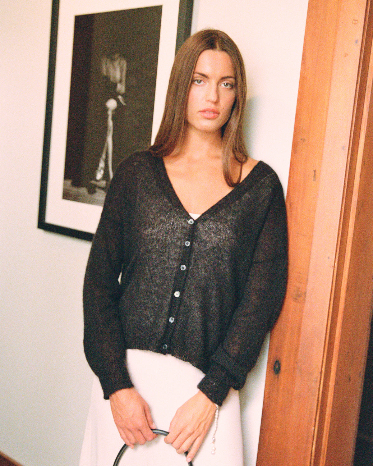 A petite woman with long brown hair stands indoors by a wooden panel, wearing the Mist Cardigan – Black and a light skirt, holding a black handle with both hands. A framed photo hangs behind her.