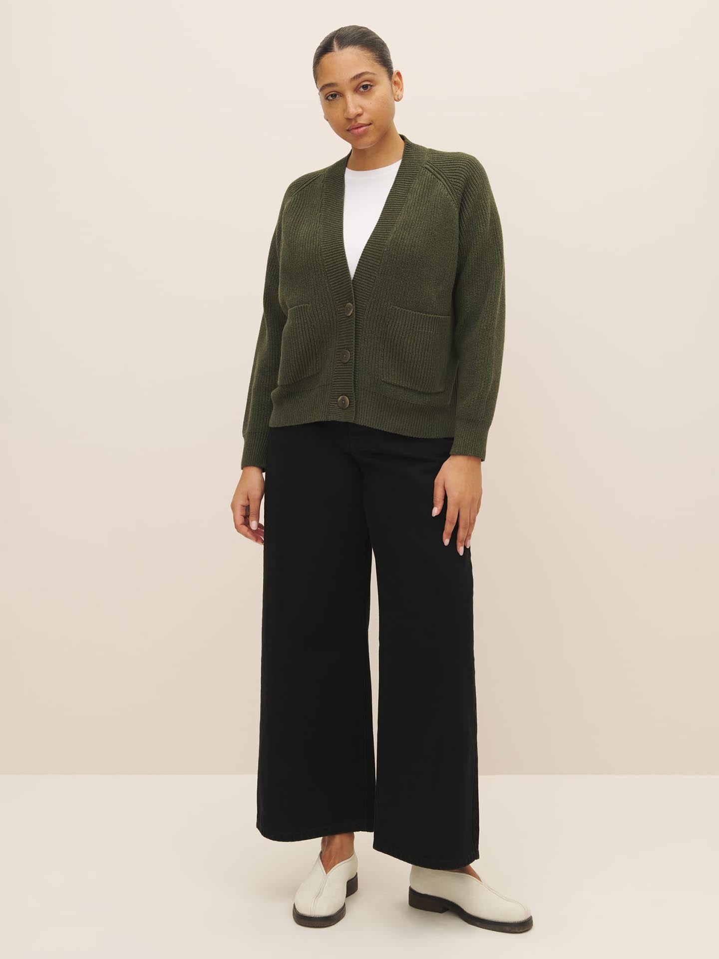 Woman posing in a Kowtow Hannes Cardigan – Khaki Marle, white top, black trousers, and white shoes against a neutral background.