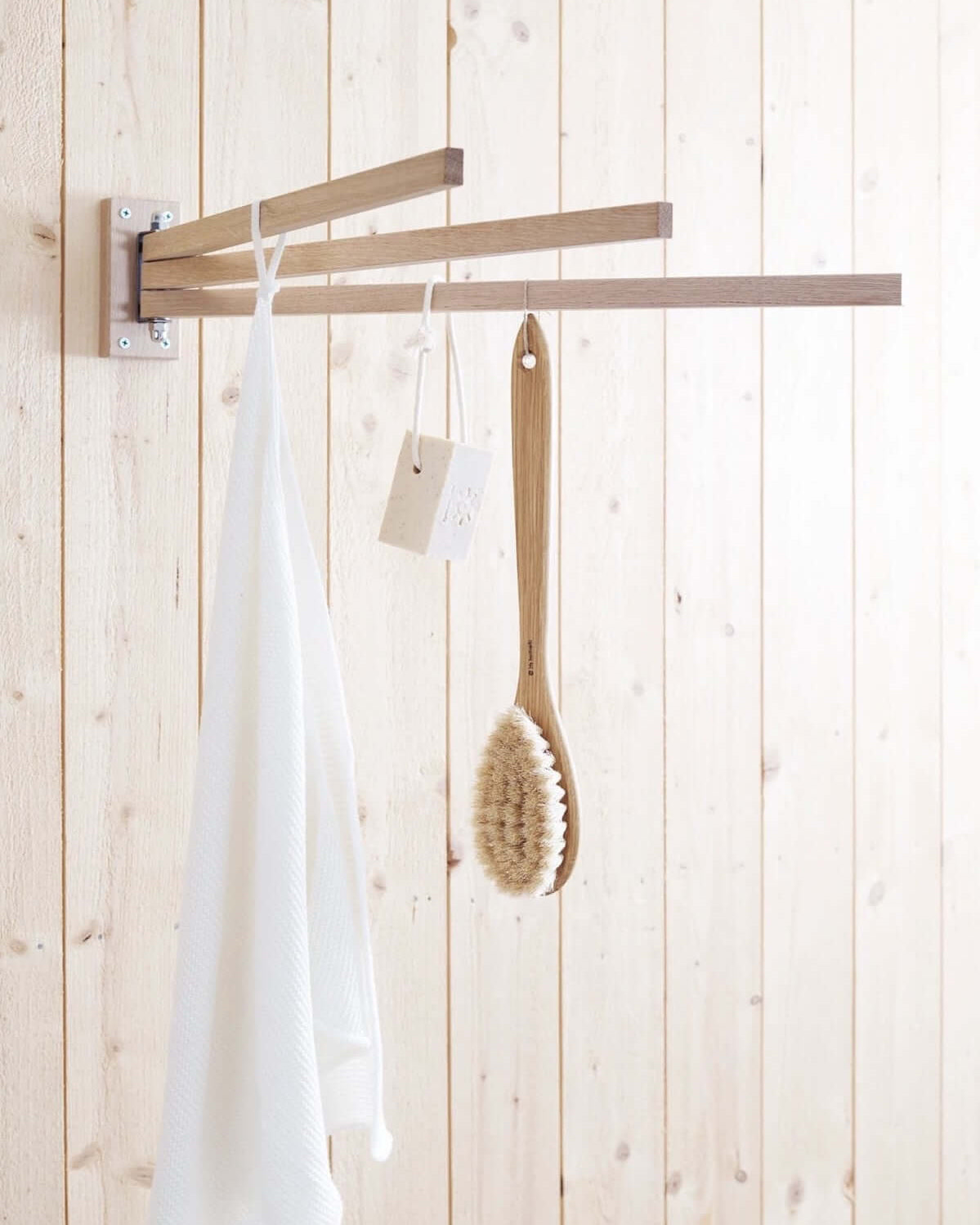 The Oak Towel Drying Rack – 3 Arm by Iris Hantverk, mounted on a light wood-paneled wall, holds a white towel, a hanging soap bar, and an Iris Hantverk wooden bath brush.