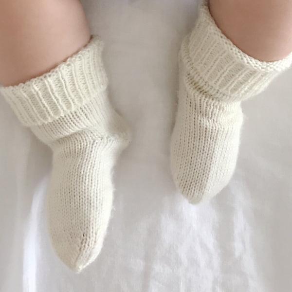 A baby&#39;s feet in Weebits Hand Knitted 2ply Merino Socks - Ivory on a bed.