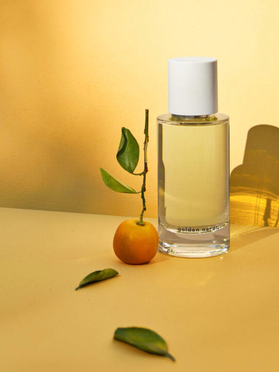 Glass perfume bottle of Golden Neroli eau de parfum by Abel next to a small citrus fruit with leaves on a warm-toned surface.