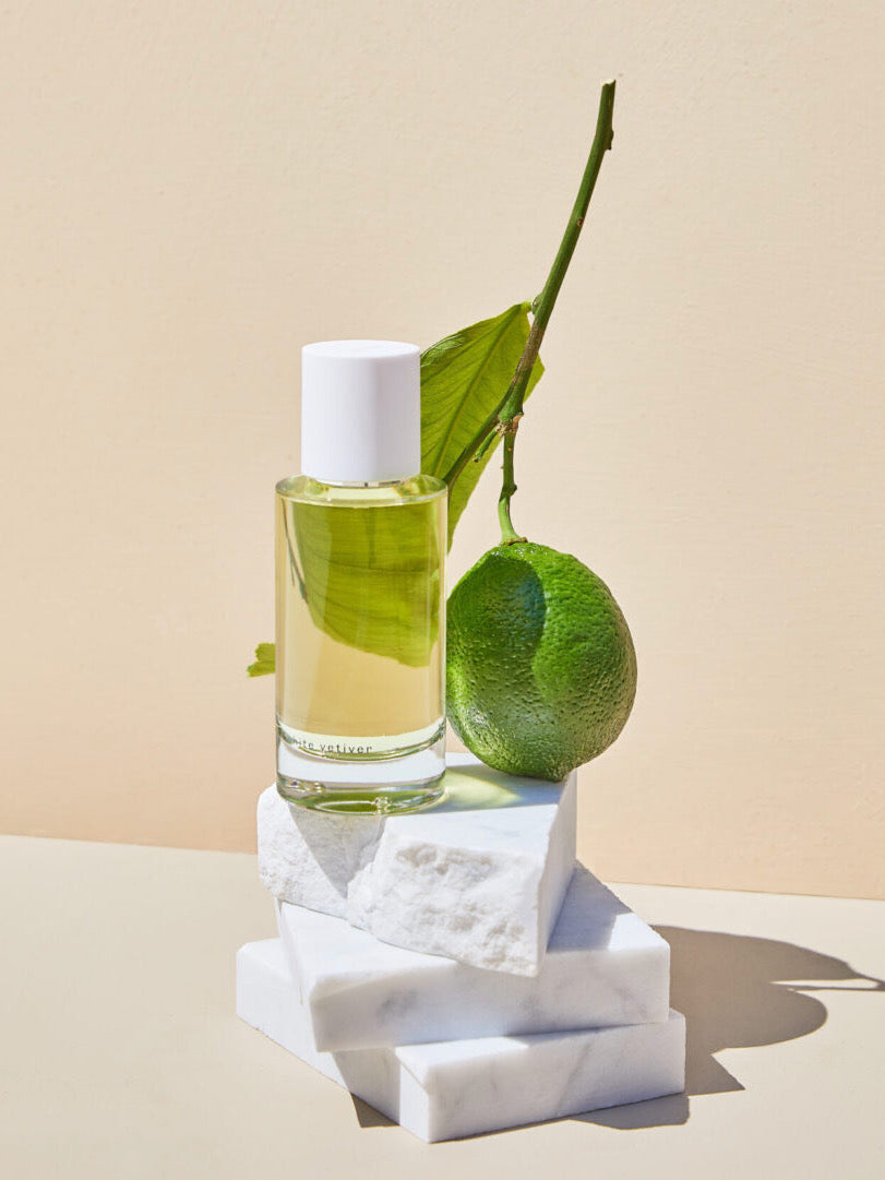 Bottle of White Vetiver eau de parfum by Abel on white marble blocks beside a green leaf and lime against a cream background.