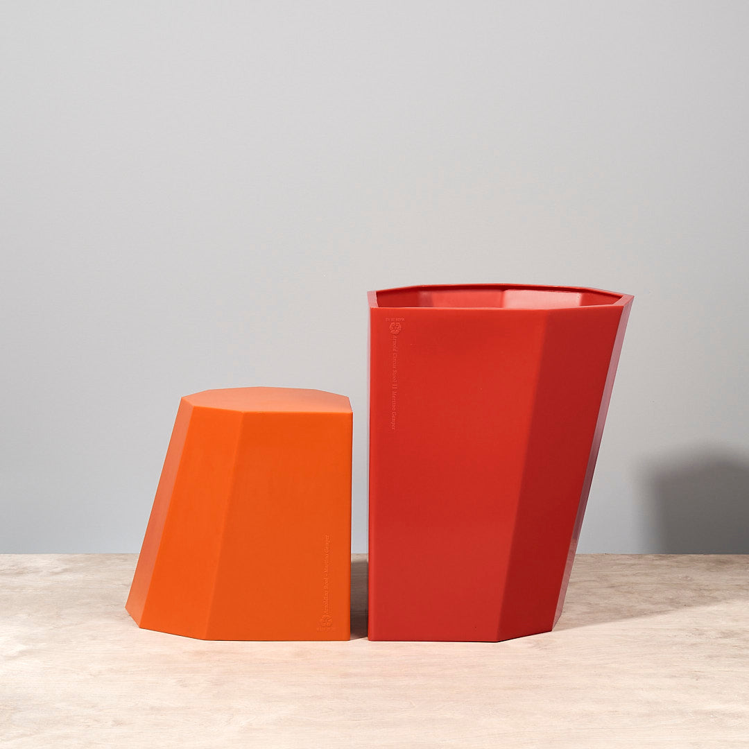 An orange vase and a Martino Gamper Arnold Circus Stool – Red on a wooden table.