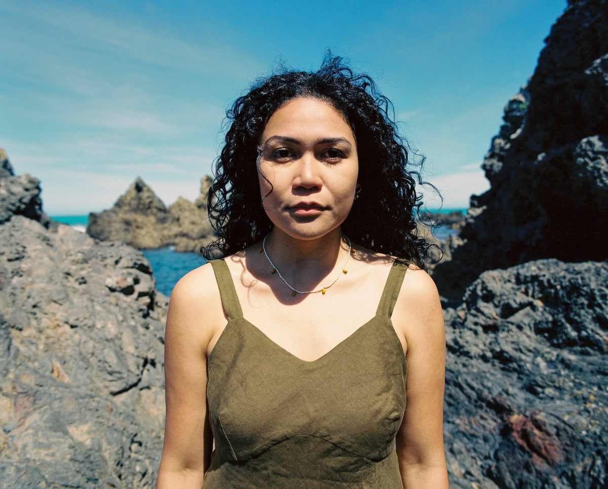 A woman in a green dress standing in front of rocks wearing the Kōwhai Seed Pearl Necklace by Avara Studio.