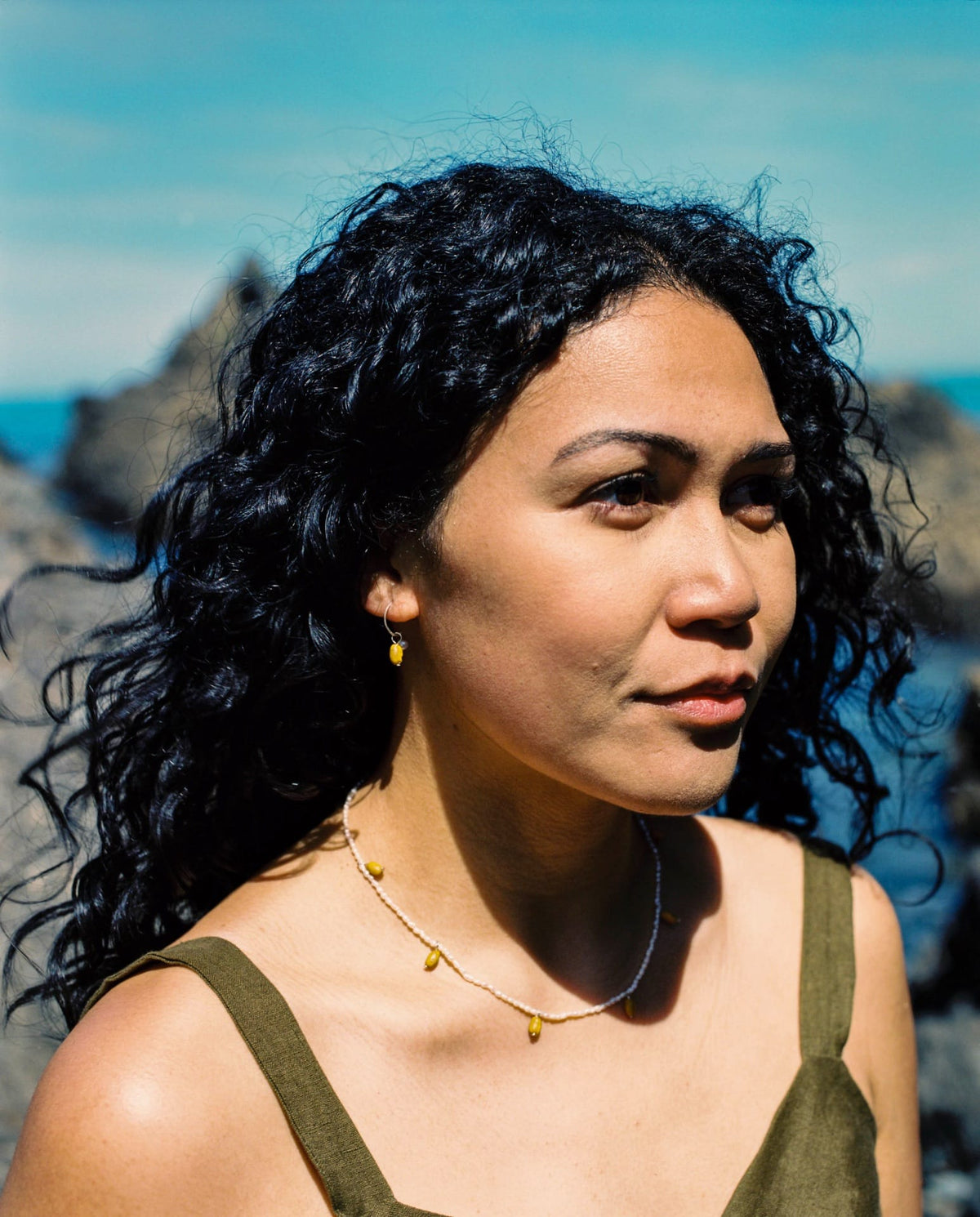 A woman with curly hair wearing Kōwhai Sleepers by Avara Studio, standing in front of the ocean.