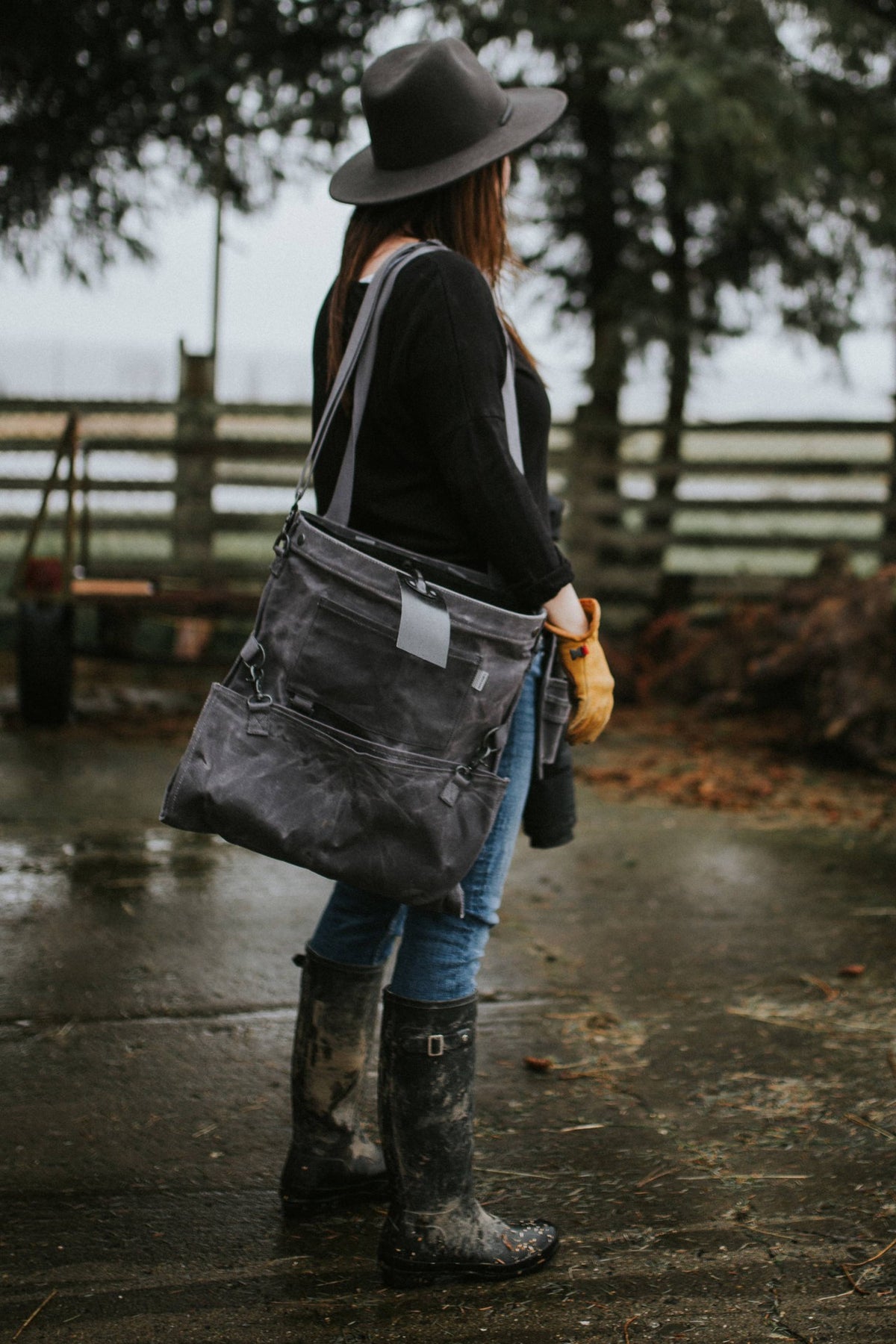 A woman wearing rain boots and a hat is holding a Barebones Gathering Bag – Grey.