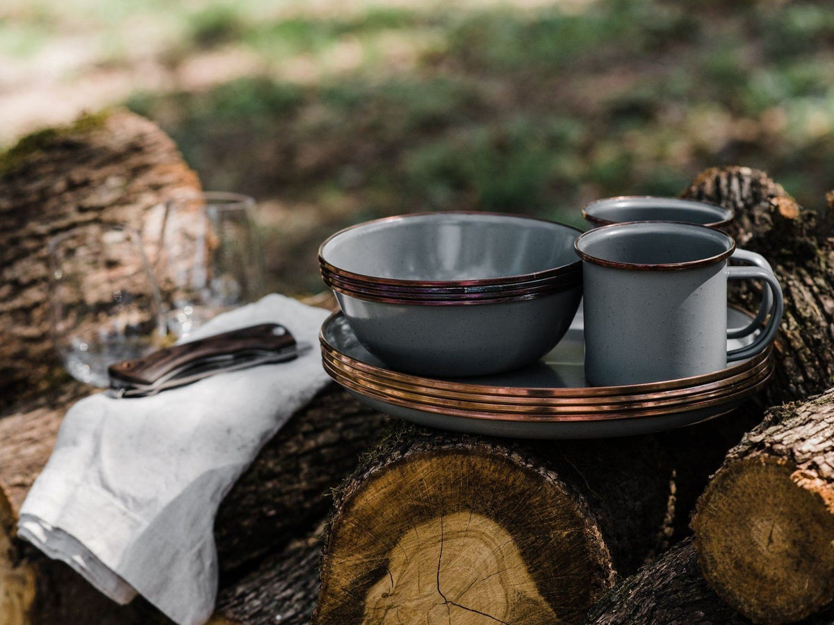 A set of Barebones enamel mugs in Slate Grey (Set of 2) on a log in a wooded area.
