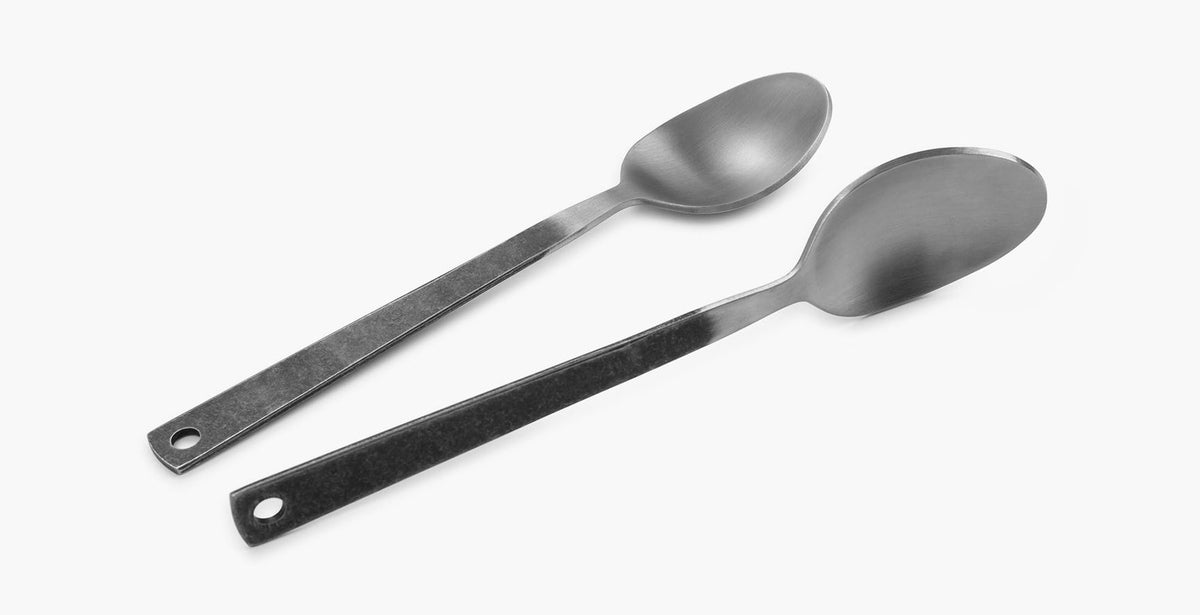 Two minimalist stainless steel spoons from Barebones&#39; Flatware Cutlery set, featuring long handles with holes at the end, are side by side on a plain white background.
