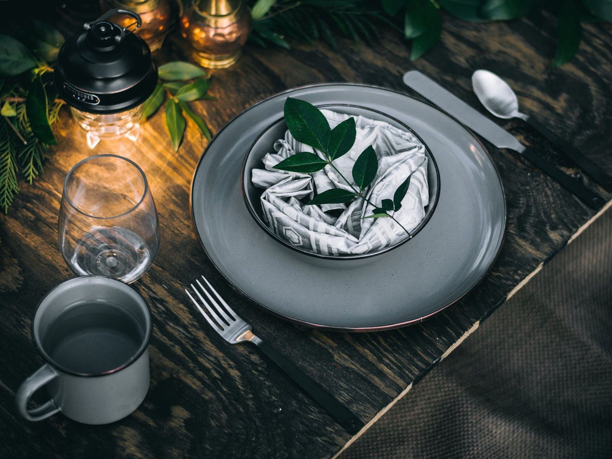 A rustic table setting features a plate, bowl with folded napkins, and green leaves. Nearby, Barebones&#39; minimalist Flatware Cutlery Set of 2 adds elegance alongside a glass, mug, spoon, knife, and lanterns on the wooden surface.