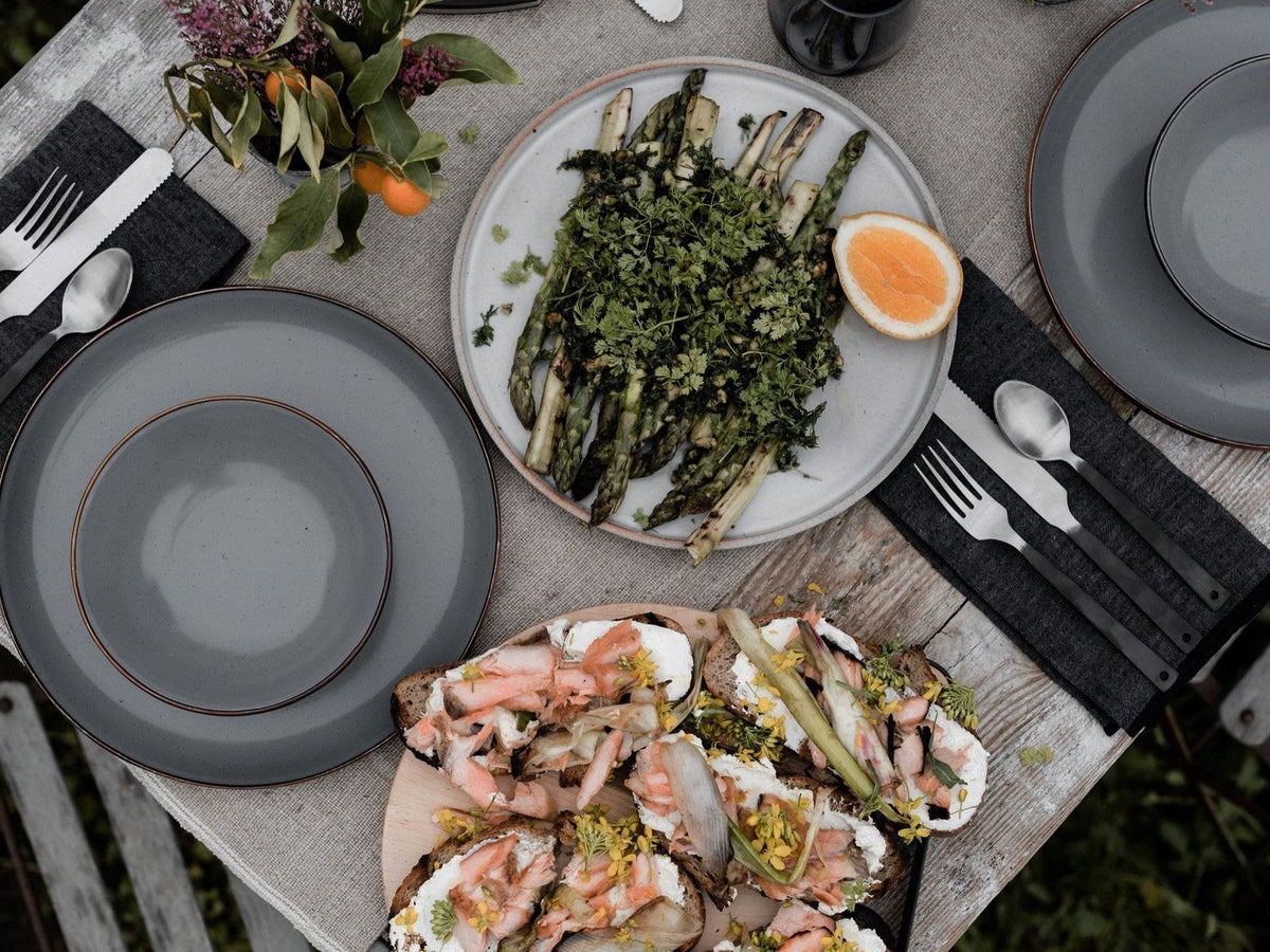 A minimalist table setting includes a Barebones Flatware Cutlery set (two forks, knives &amp; spoons) and two empty plates. A floral centerpiece adds charm alongside asparagus with herbs and open-faced salmon sandwiches.