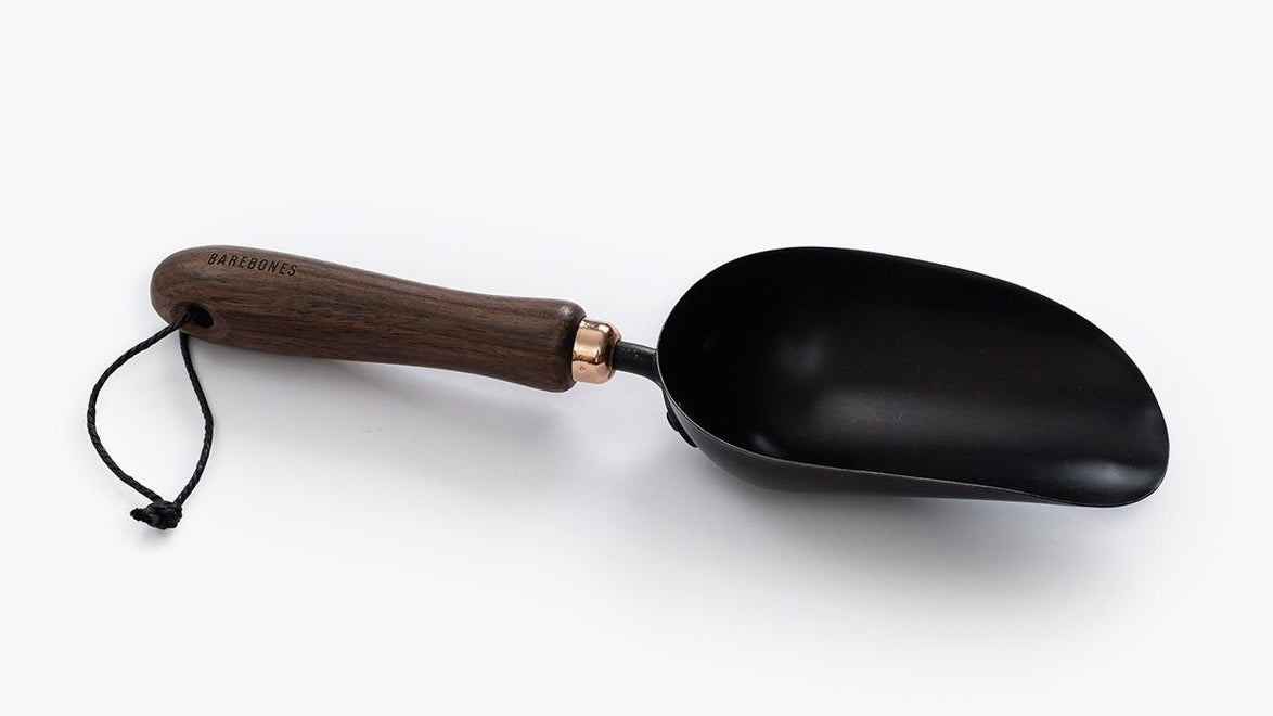 A black Garden Scoop with a wooden handle on a white surface, by Barebones.