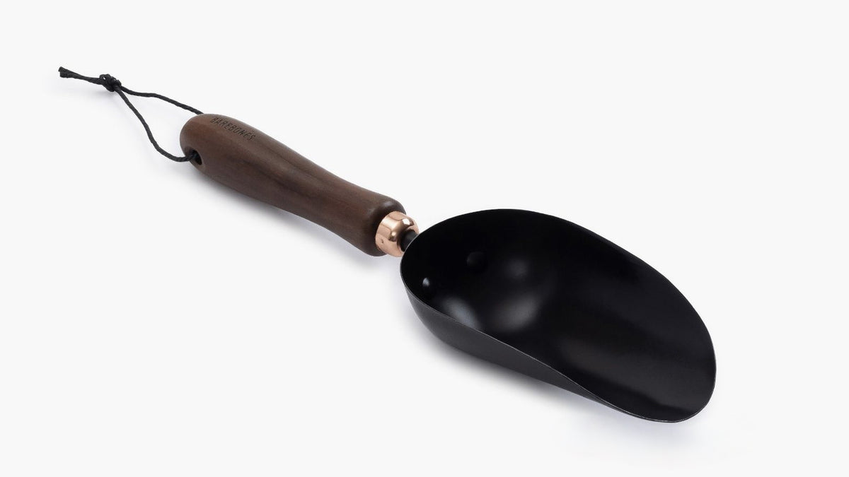 A black Garden Scoop with a wooden handle on a white surface by Barebones.