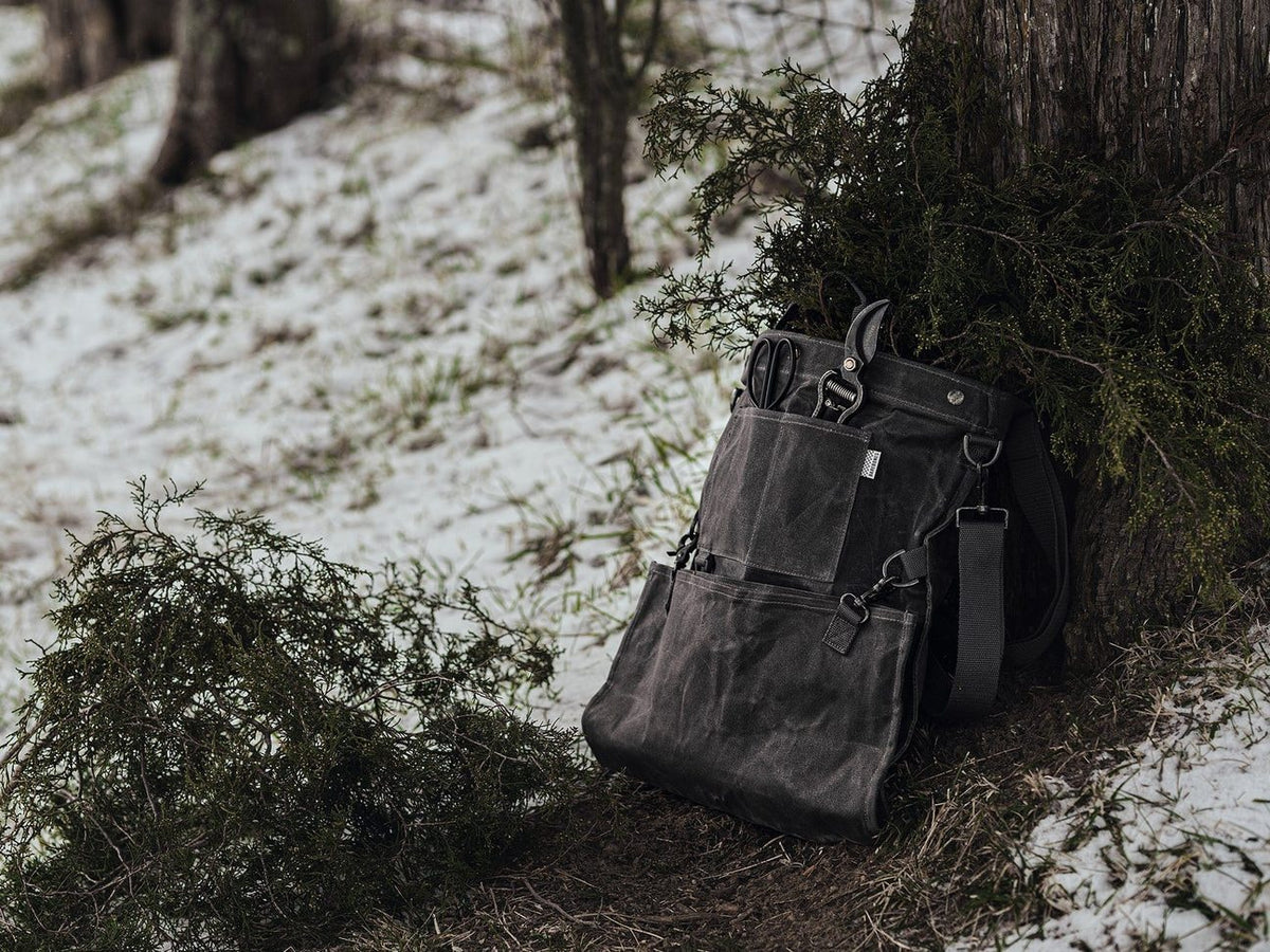 A Gathering Bag – Grey backpack leaning against a tree in the snow, Barebones.