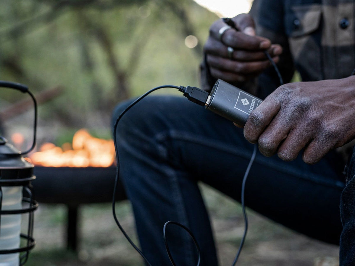A man is using a Barebones Portable Charger – Integrated Torch in front of a campfire.