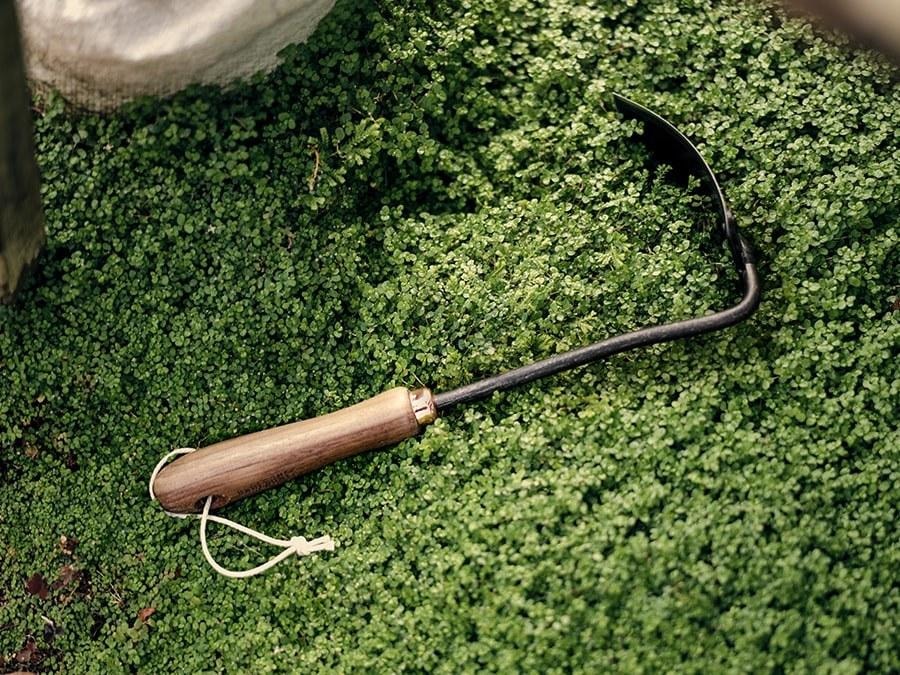 A Barebones Square Hand Hoe is laying on the grass next to a bag.