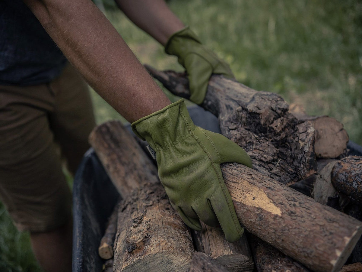 A man wearing Barebones Classic Work Gloves – Olive is putting wood in a wheelbarrow.