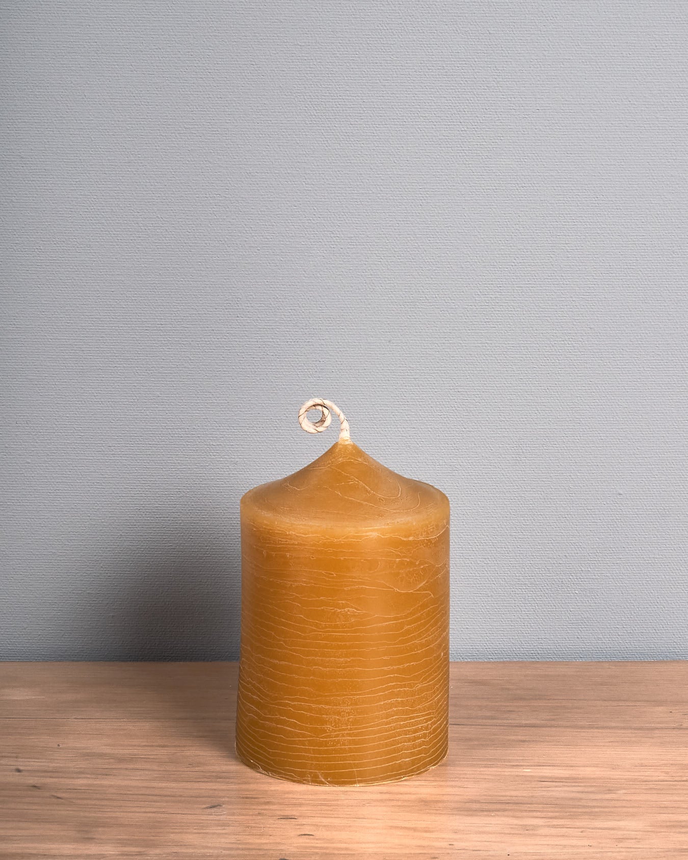 A tall Pillar Beeswax Candle – Fat, handcrafted by Hohepa Candles, with a spiral wick rests on a wooden surface against a plain gray wall.
