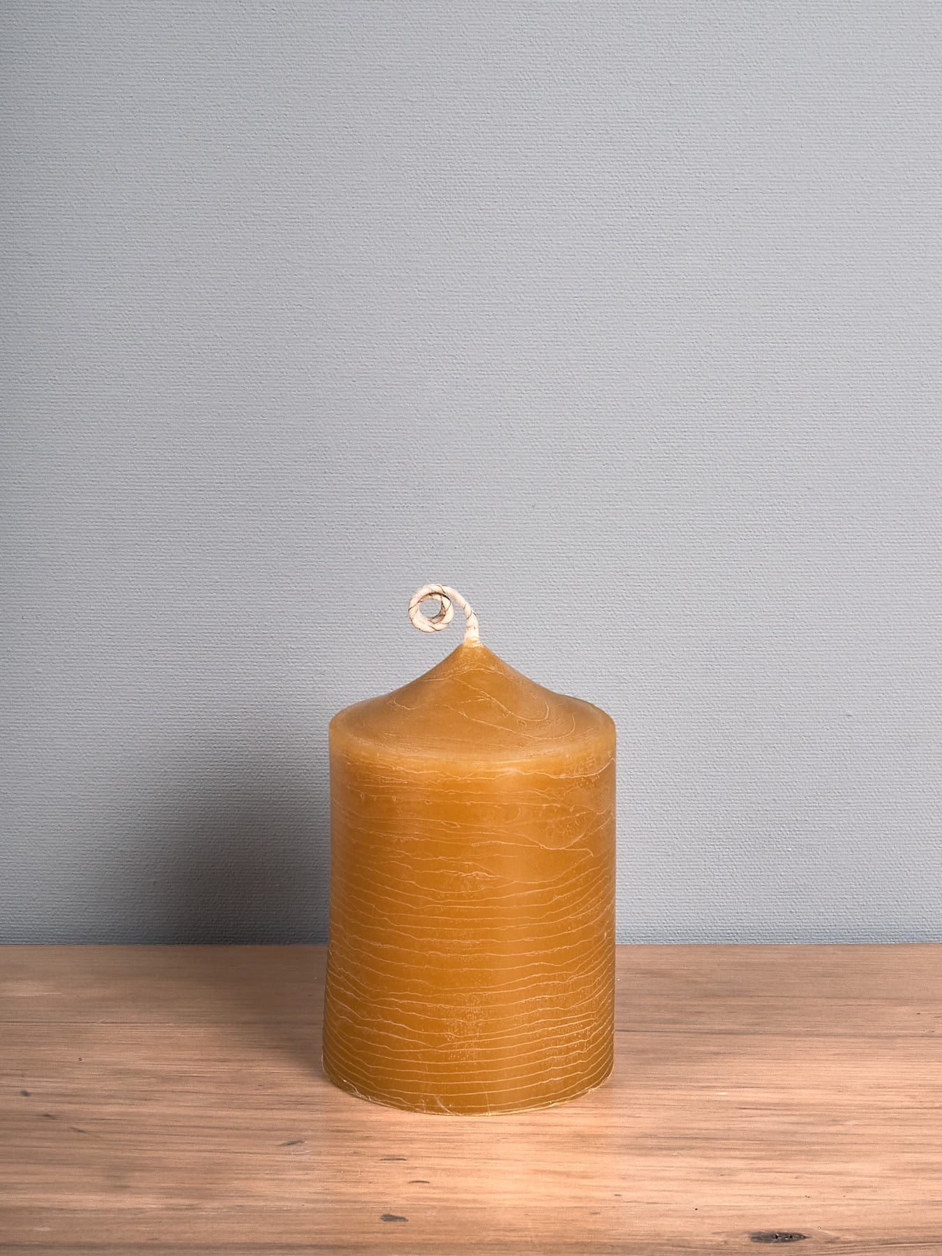A tall Pillar Beeswax Candle – Fat, handcrafted by Hohepa Candles, with a spiral wick rests on a wooden surface against a plain gray wall.