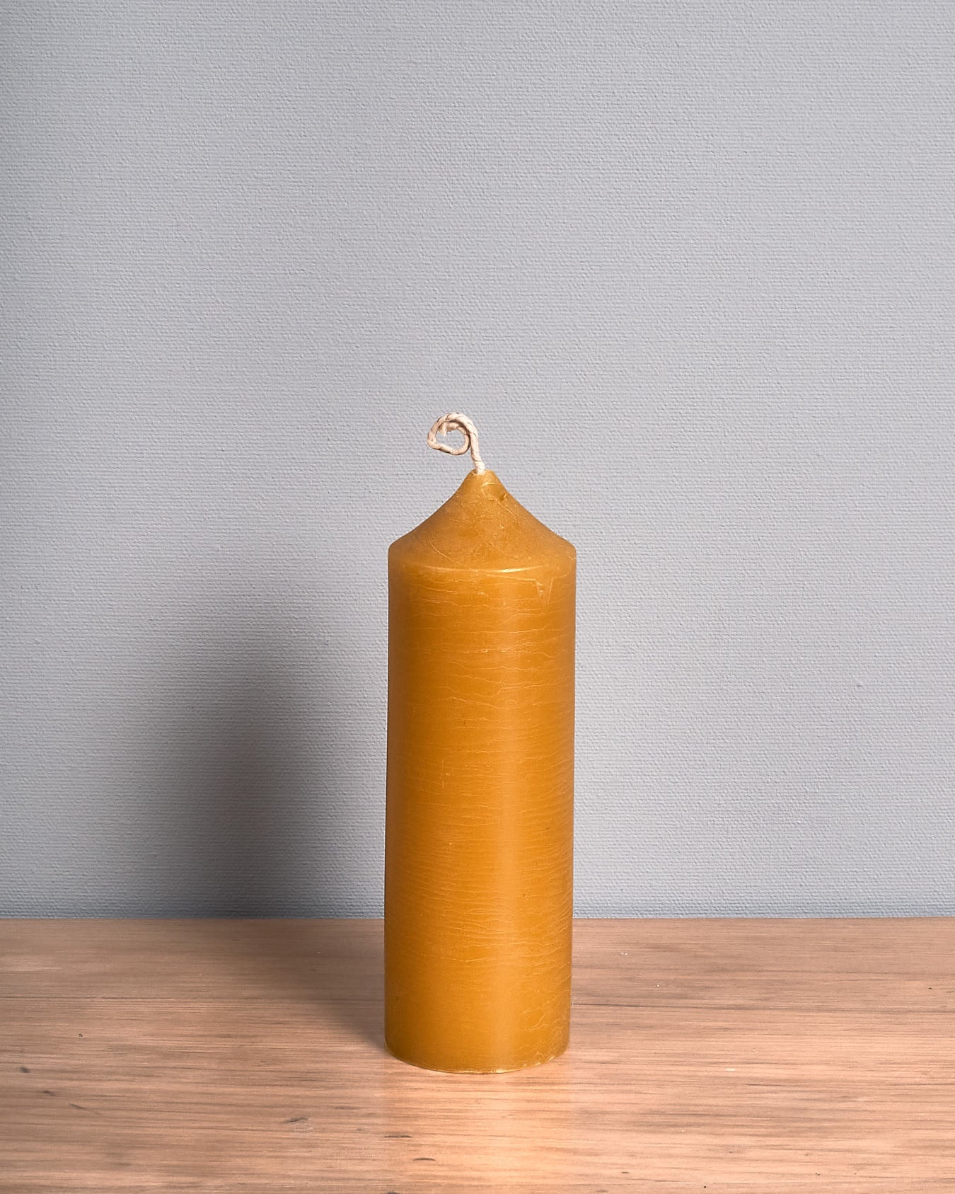 The Pillar Beeswax Candle – Medium by Hohepa Candles, featuring a tall cylindrical shape and a pointed top, in its natural yellow hue, is positioned on a wooden surface against a gray background.