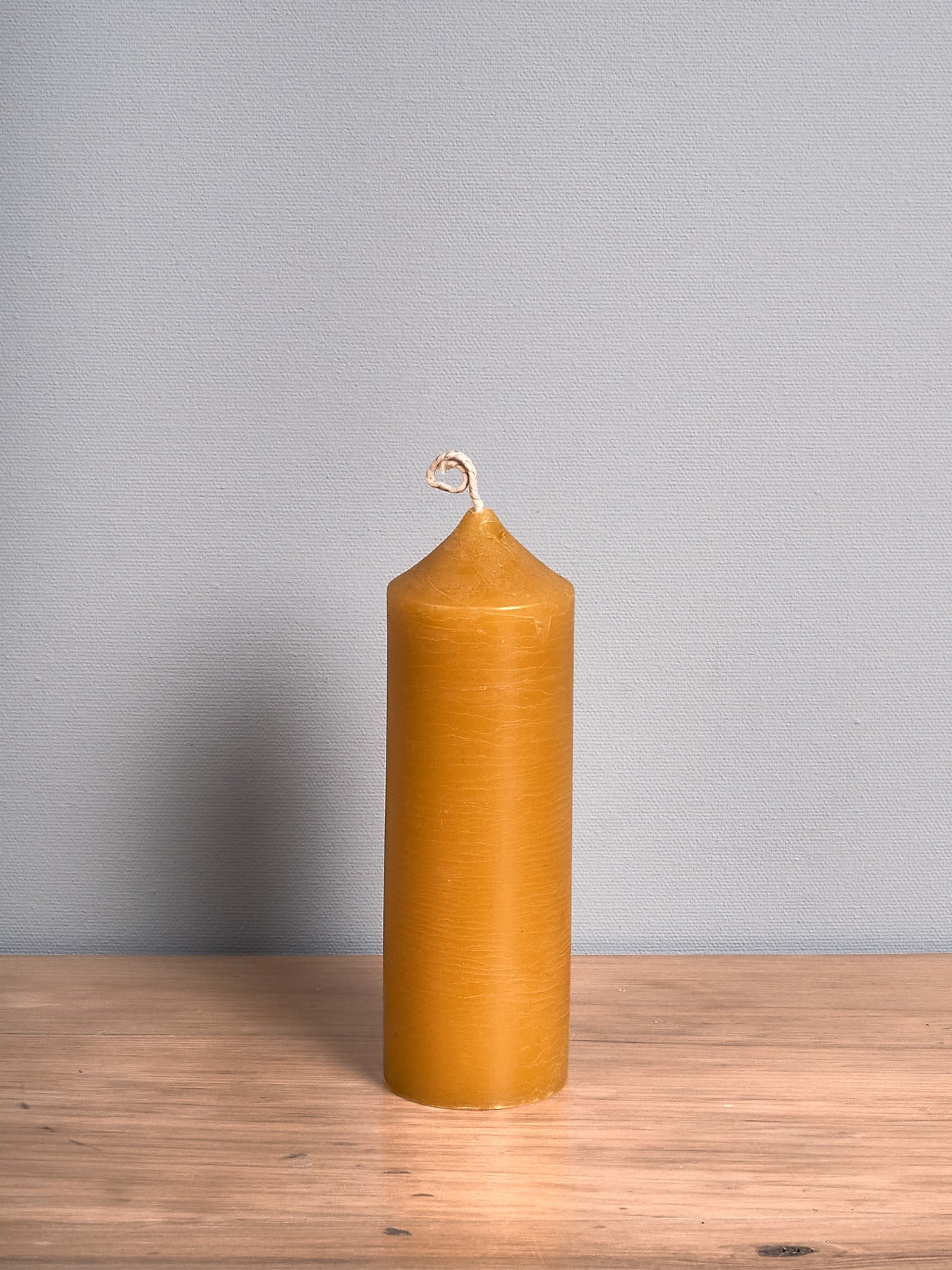 The Pillar Beeswax Candle – Medium by Hohepa Candles, featuring a tall cylindrical shape and a pointed top, in its natural yellow hue, is positioned on a wooden surface against a gray background.