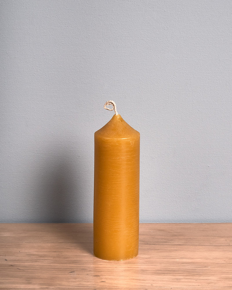 The Pillar Beeswax Candle – Medium by Hohepa Candles, featuring a tall cylindrical shape and a pointed top, in its natural yellow hue, is positioned on a wooden surface against a gray background.