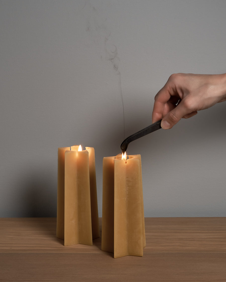 A hand is using a tool to extinguish the flame of one of three tall, Hohepa Candles Star Pillar Beeswax Candles on a wooden surface.