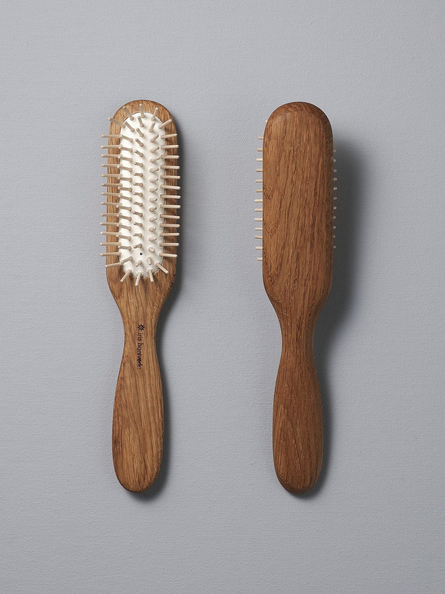 Two Iris Hantverk Oak Hairbrushes with smooth handles, one showing the bristles side with natural rubber pins and the other the back, positioned side by side on a gray background. Perfect for a soothing scalp massage.