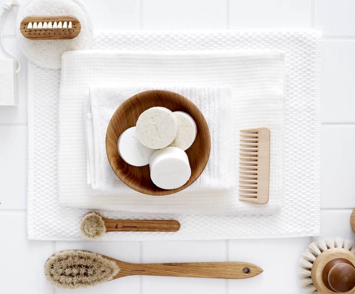 A white towel, soap, Face Brush – Dry (Iris Hantverk) and toothbrush on a tiled floor.