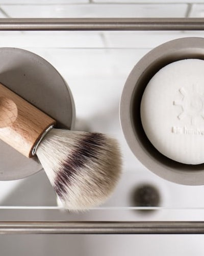 A Shaving Brush – Silver Tip by Iris Hantverk with a wooden handle and cruelty-free synthetic fibres stands upright on a light gray surface against a light gray background.