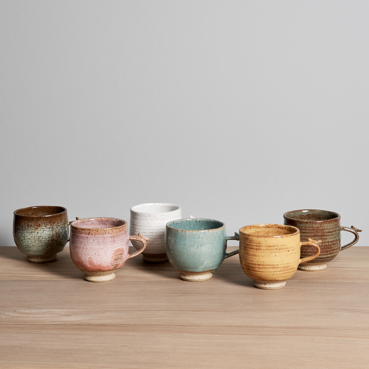 Five different colored Bird Handle Cups - Rhubarb are lined up on a wooden table, made by Jino Ceramic Studio.