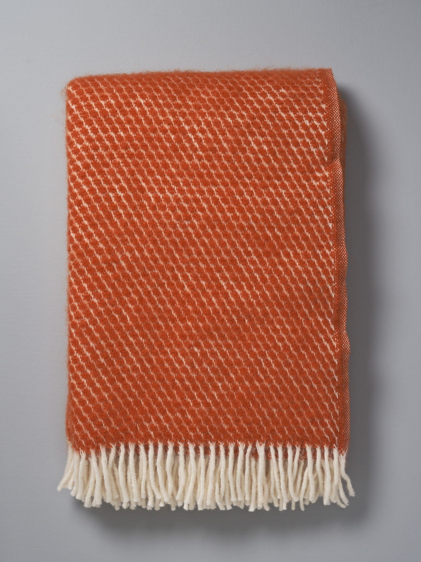 A Honeycomb ECO* Lambswool Throw - Rust by Klippan with white fringes on a grey background.